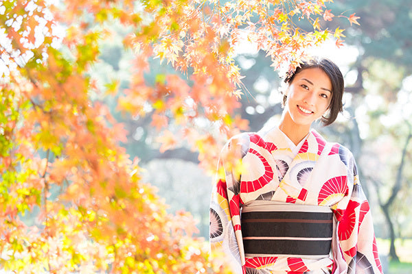 A lady in a red and white kimono stands under vibrant autumn leaves in a sunny garden