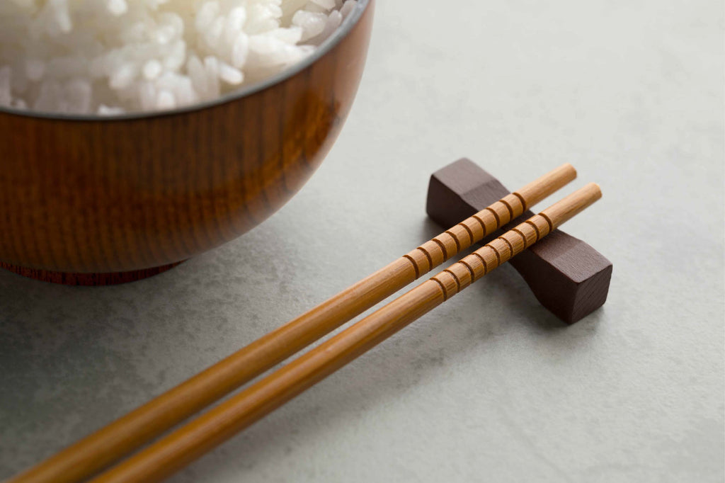 A wooden bowl brimming with white rice, with chopsticks on a dark holder