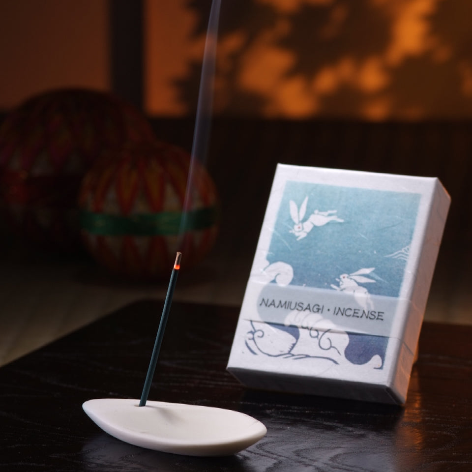 A burning incense stick on a white holder next to a namusagi incense box