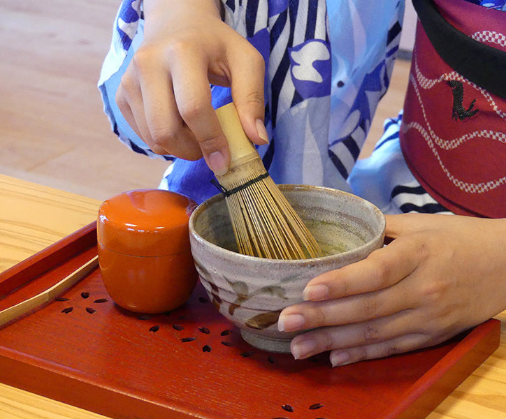 An individual in Japanese attire stirring matcha tea in a ceramic bowl