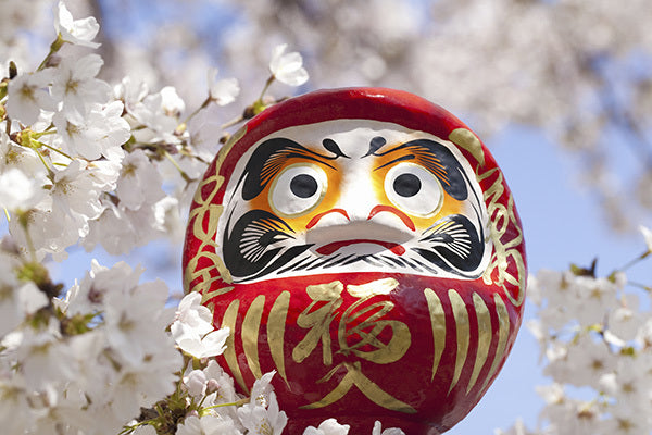 A vivid red daruma doll peers through white cherry blossoms against a clear blue sky