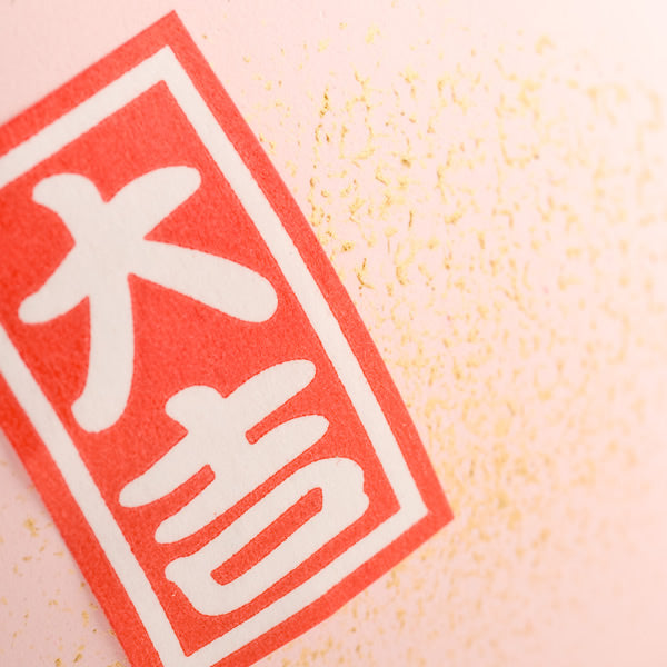 Close-up of a red seal with Japanese characters on a pink, gold-speckled textured backdrop.